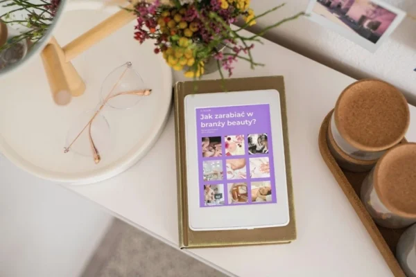 Okładka e-booka „Jak zarabiać w branży beauty” wyświetlona na tablecie leżącym na biurku obok okularów.